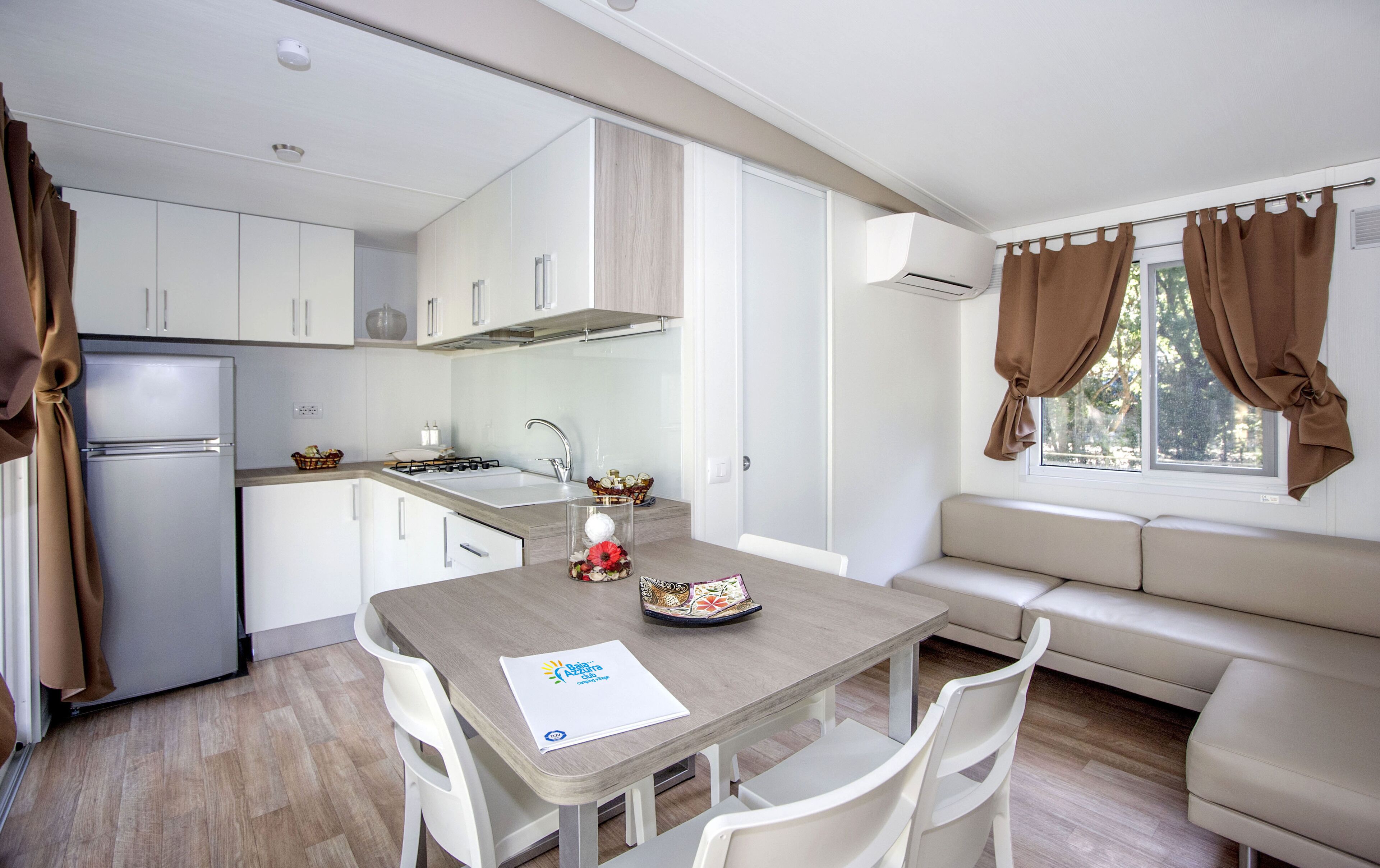 A compact kitchen with white cabinets, a refrigerator, a stove, and a sink. There's a dining table with chairs and a sofa near a window with brown curtains.