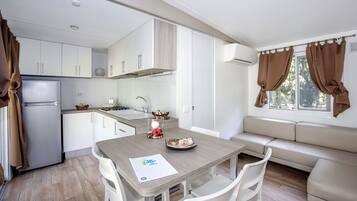 A compact kitchen with white cabinets, a refrigerator, a stove, and a sink. There's a dining table with chairs and a sofa near a window with brown curtains.