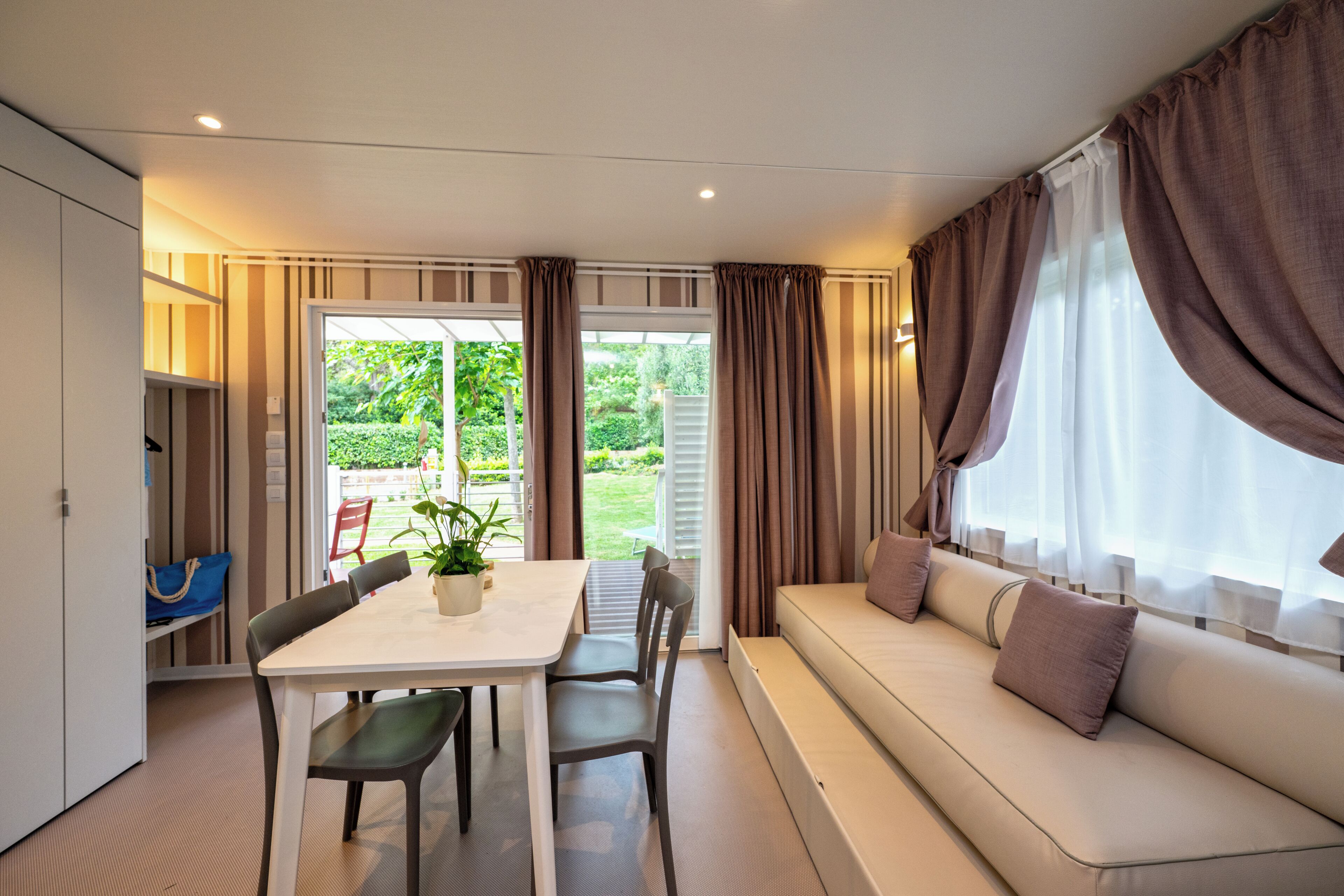A modern room with a dining table, chairs, a sofa, and a view of a garden through a glass door.