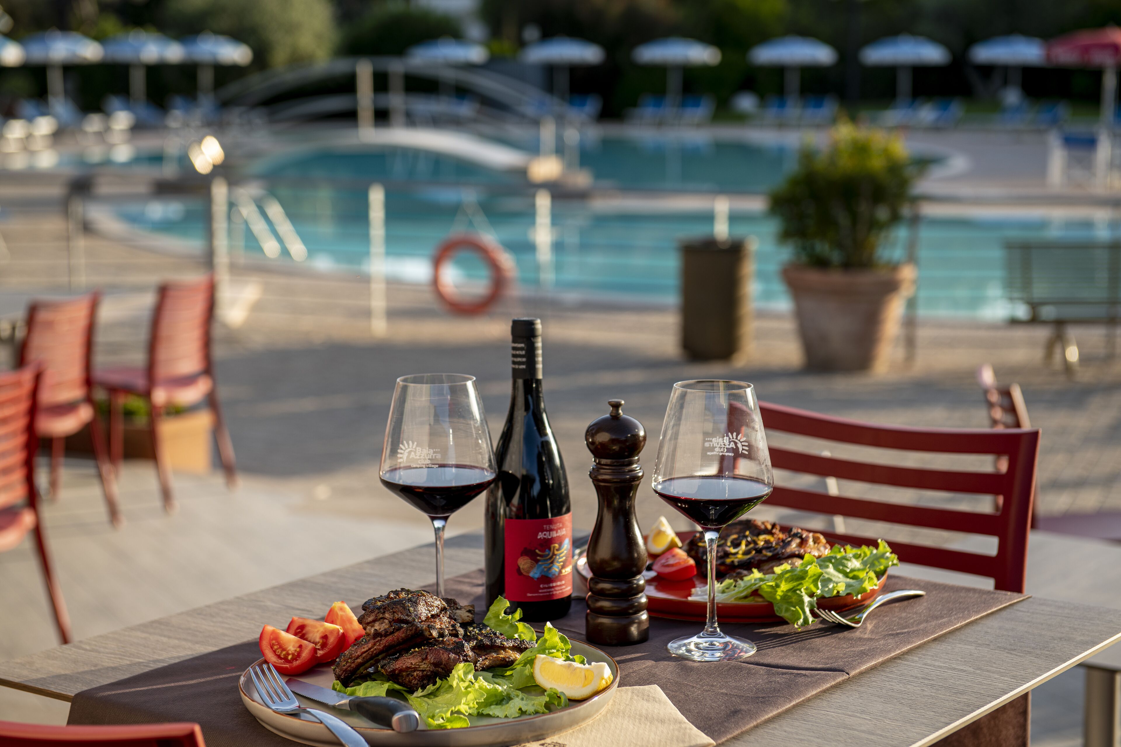 Ein Esstisch mit Mahlzeit, Weingläsern und einer Flasche Aquamarine Wein, vor einer Poolanlage.