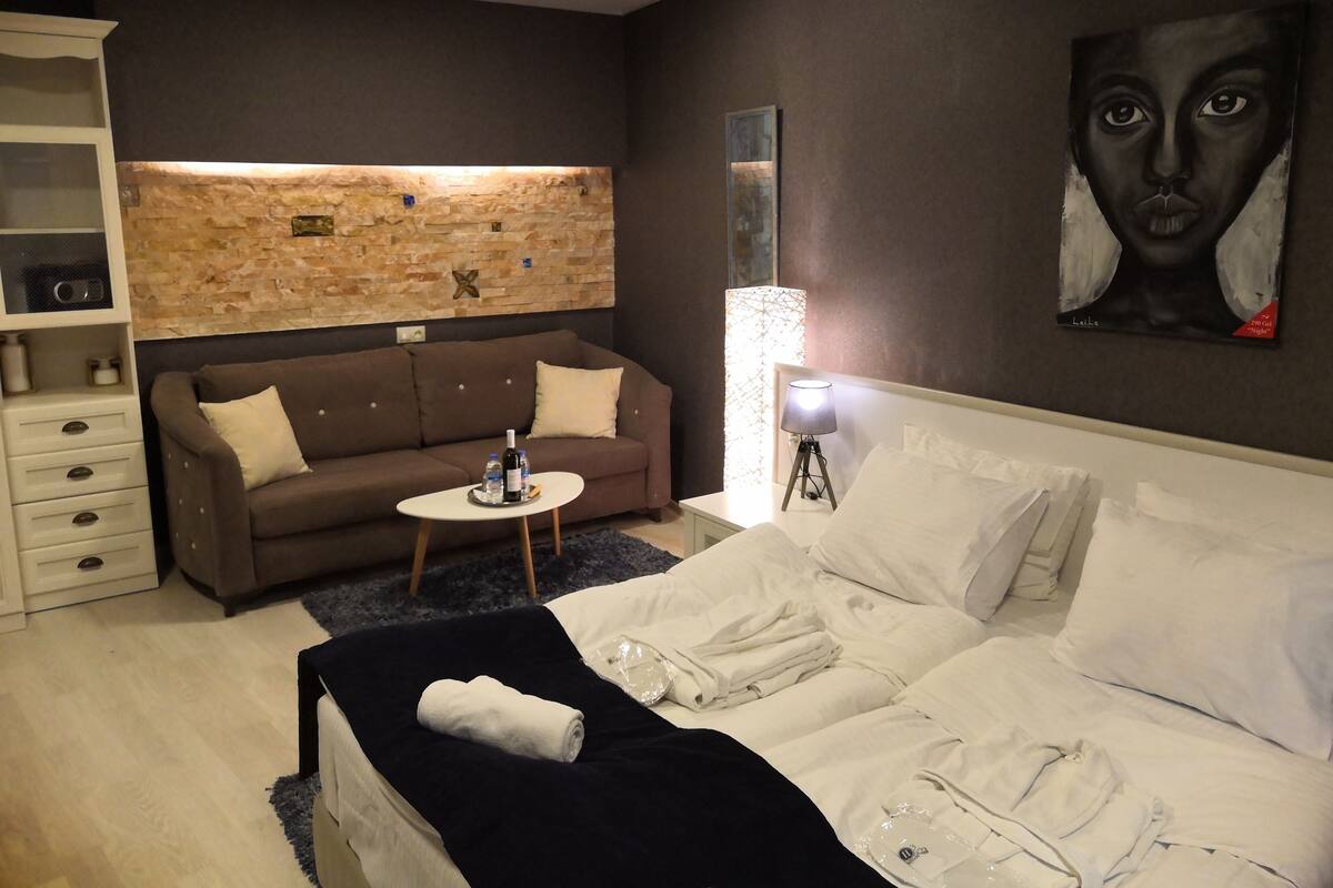 A modern bedroom with a bed, a sofa, a small table with a bottle and water, a lamp, and a painting on the wall.