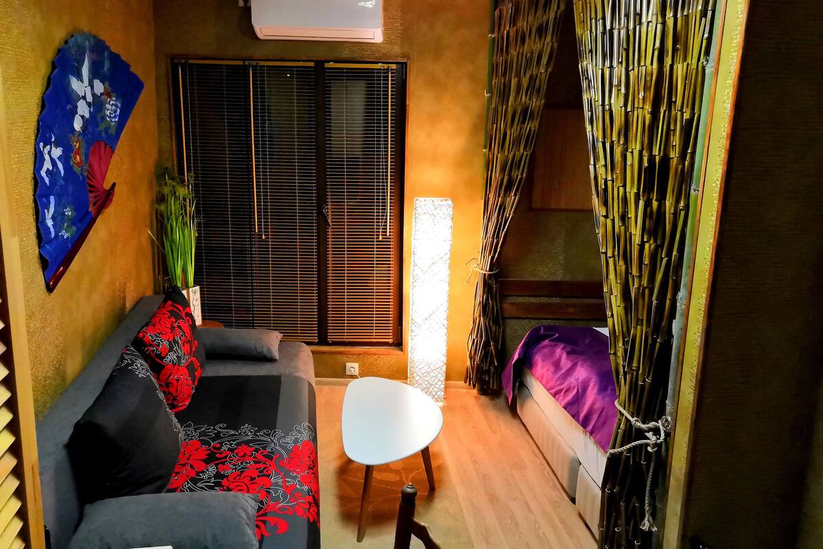 A compact living space with a sofa, a small table, a window with bamboo curtains, and an air conditioning unit.