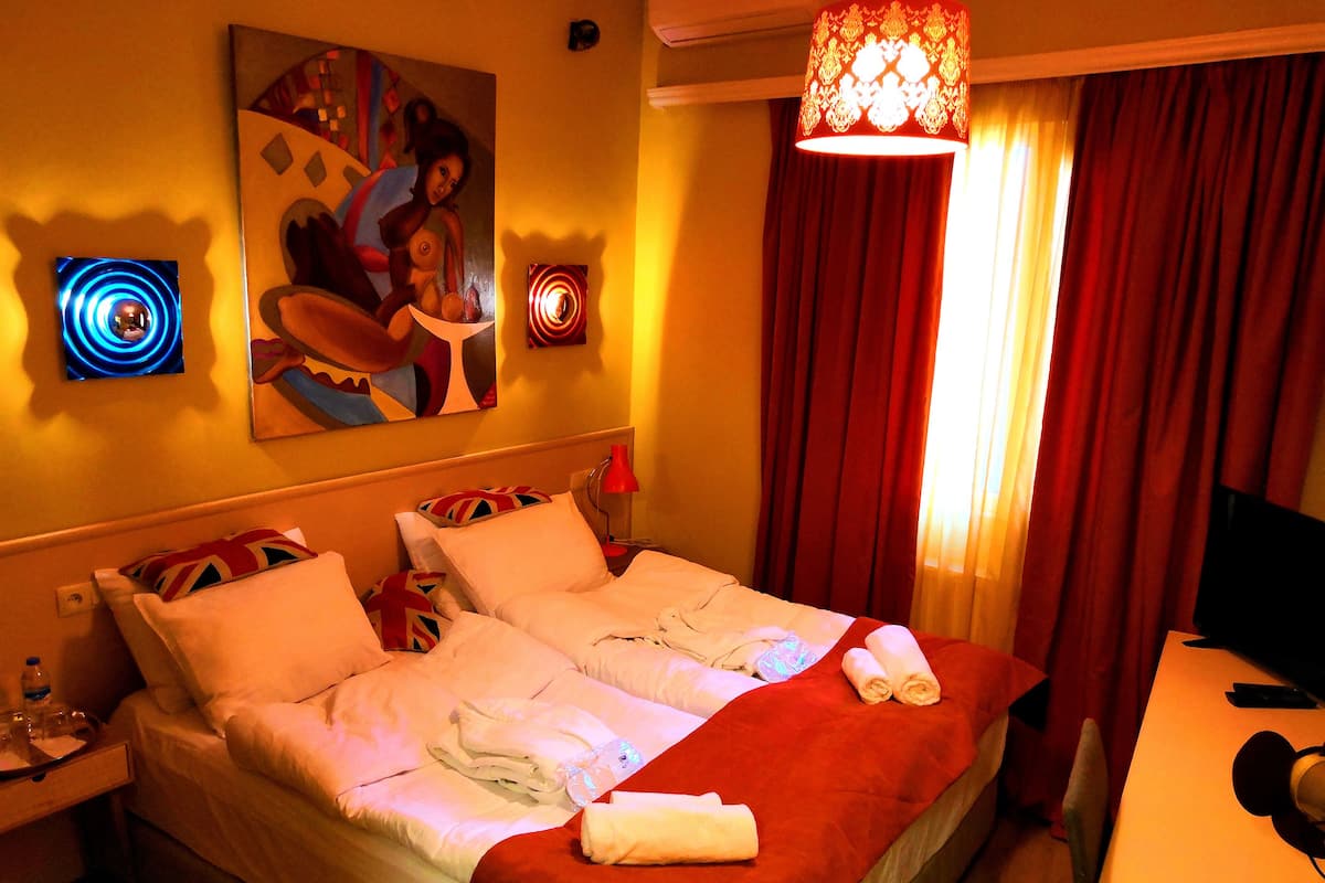 A hotel room with a double bed, red curtains, a painting on the wall, and a desk with a computer.
