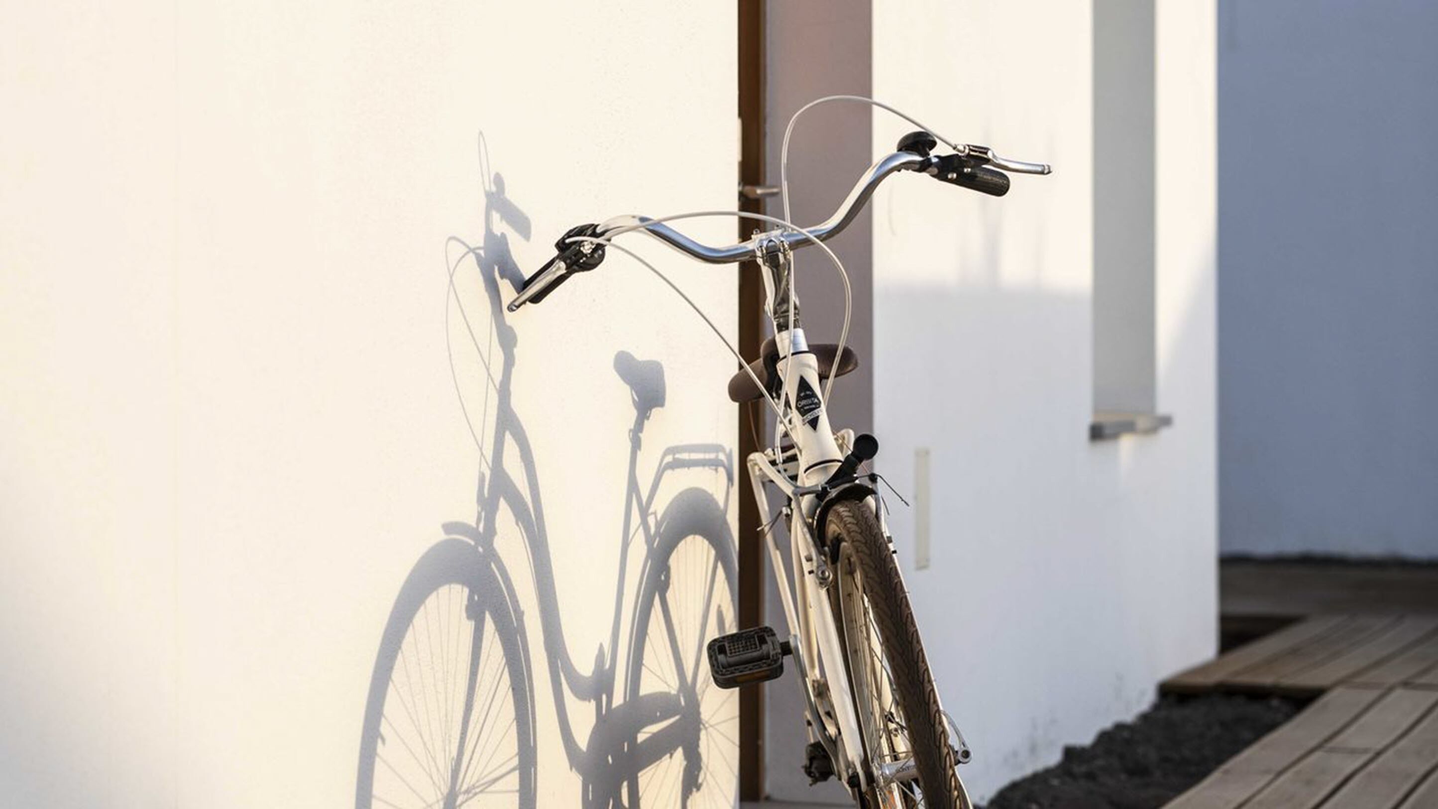 Een fiets leunt tegen een witte muur, met de schaduw van een andere fiets.