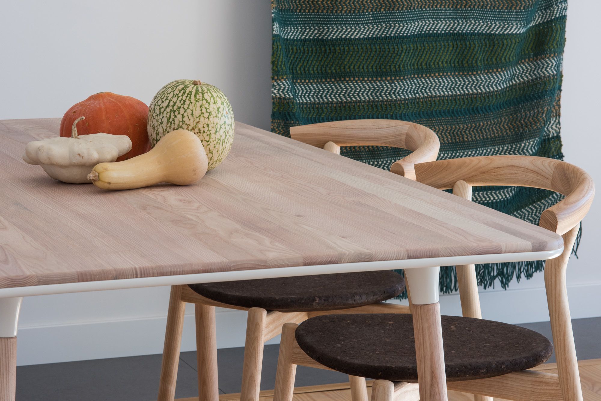 A dining table with a light wood finish, accompanied by a green woven chair and a dark wooden stool.