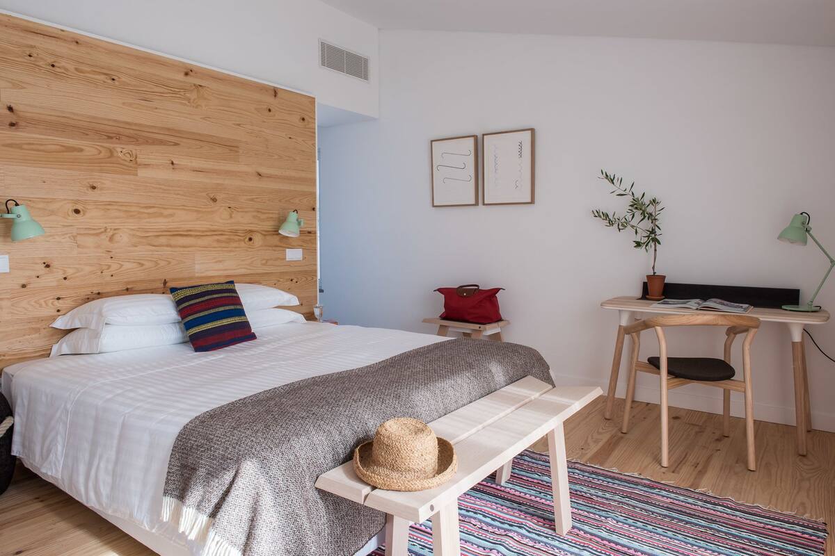 A bedroom with a wooden headboard, a bed with white bedding, a bench with a straw hat, a small table with a plant, and a desk with a lamp.