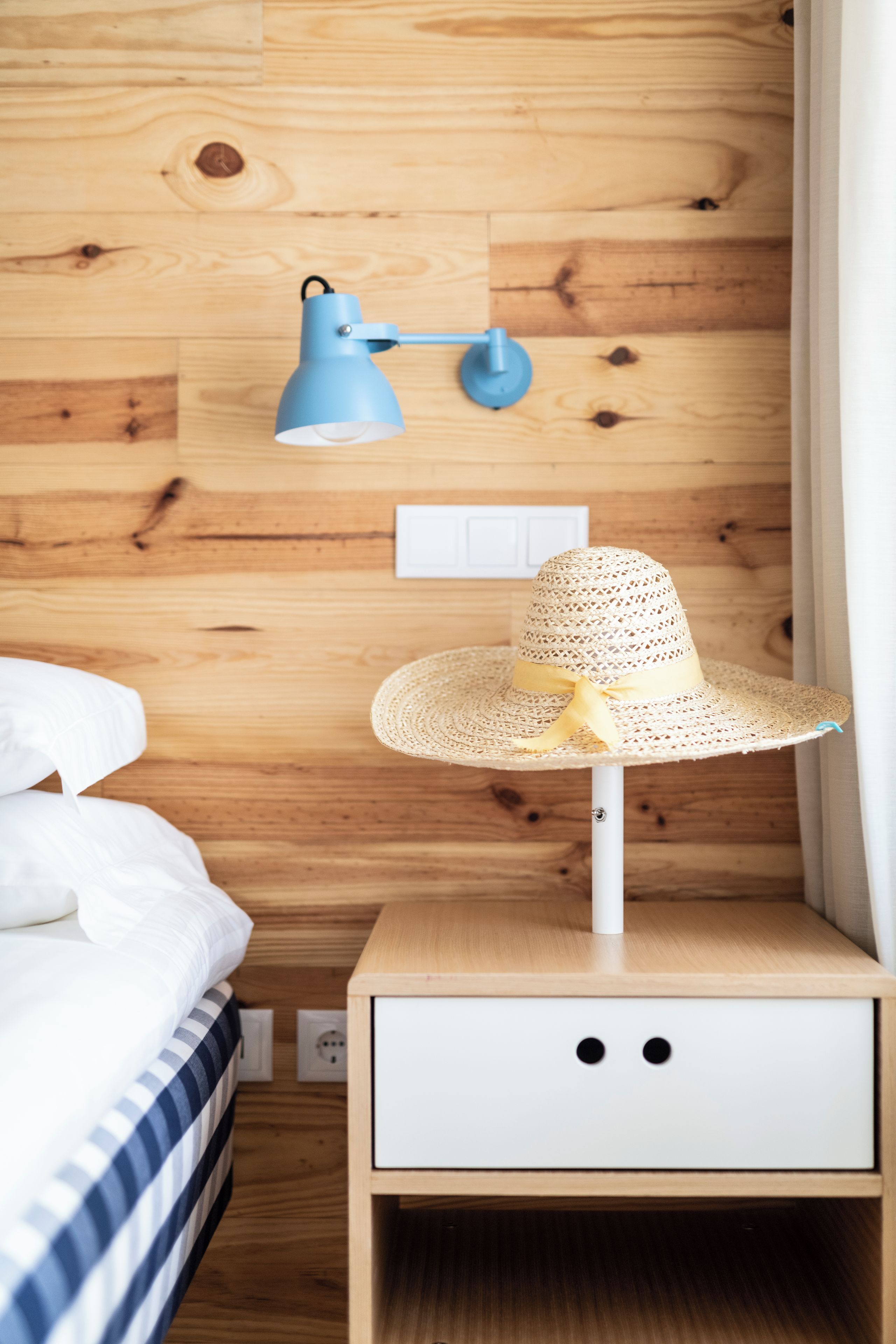 A wooden wall with a blue lamp, a white bedside table with a drawer, and a straw hat on the table.