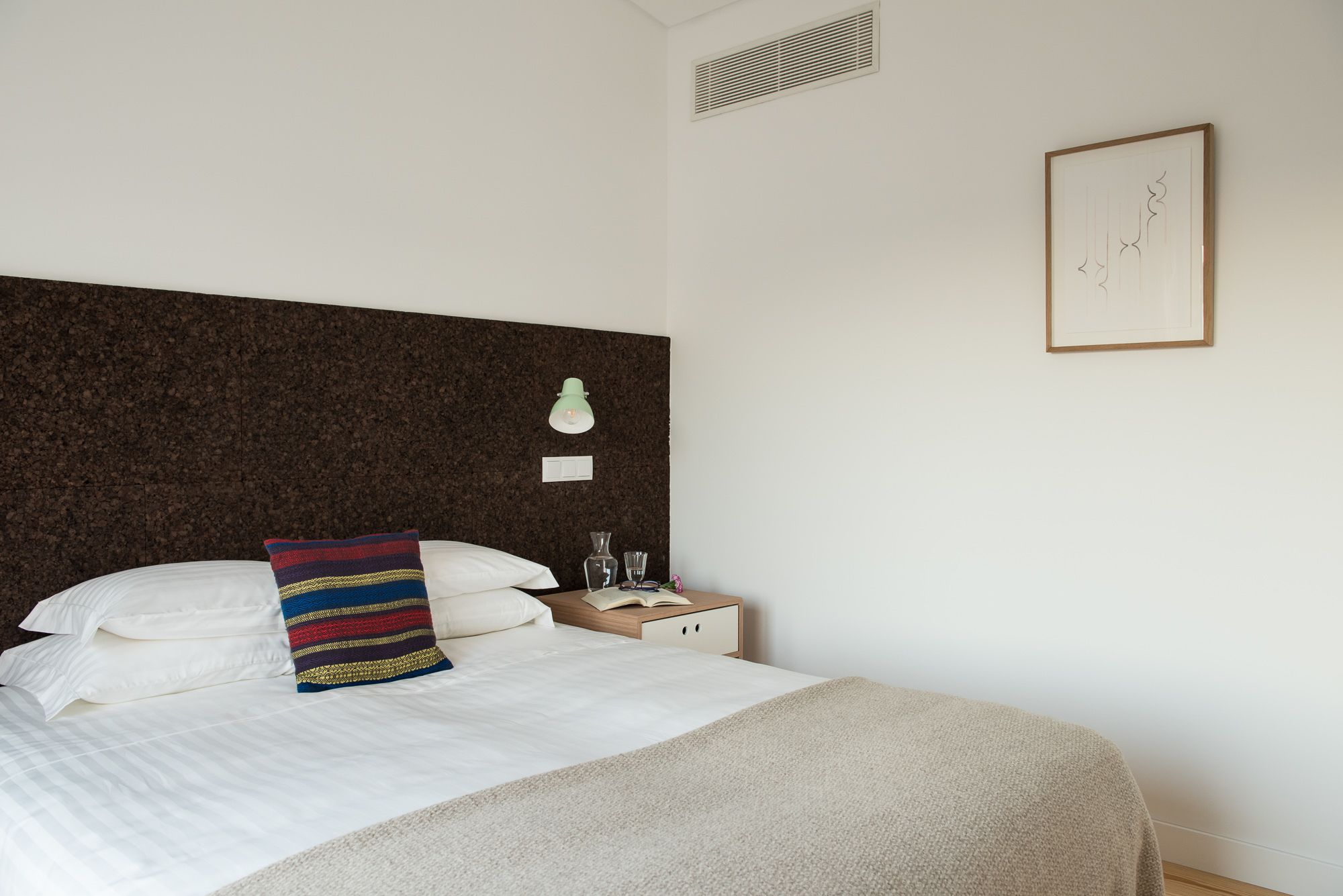 A neatly made bed with white linens and a colorful striped pillow, a bedside table with a lamp, and a framed picture on the wall.