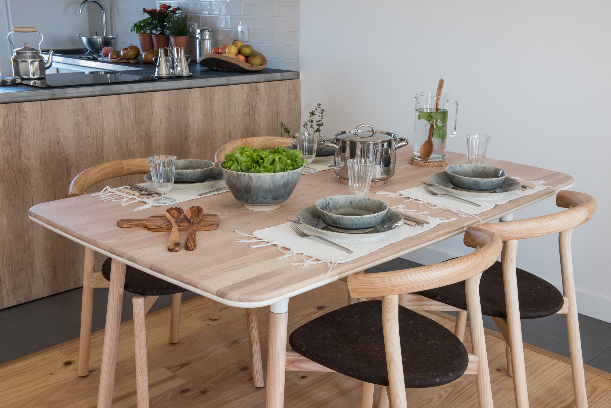 A wooden dining table with four chairs, a bowl of green salad, and various kitchen items on the counter.