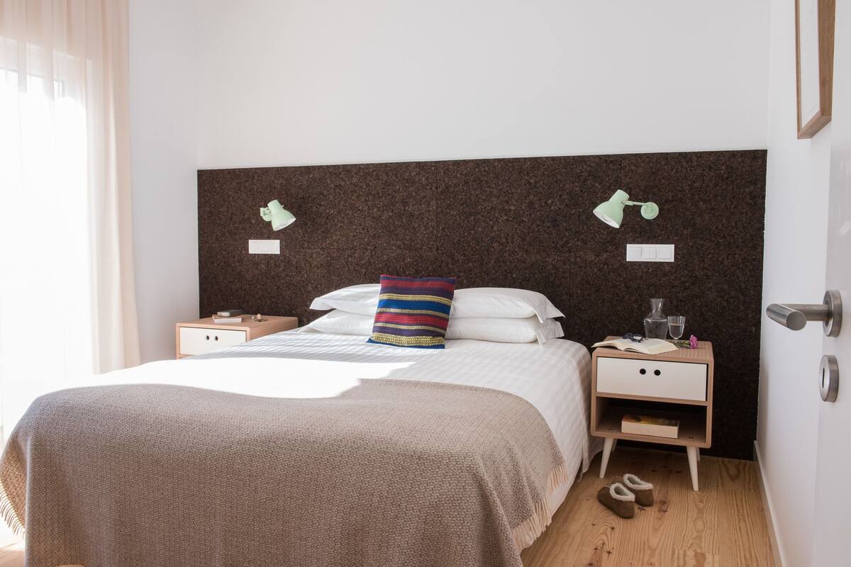 A neatly arranged bedroom with a bed, bedside tables, a nightstand, a lamp, slippers, and a door.