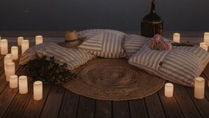 Un espace extérieur paisible, aménagé avec des coussins et des bougies, offrant une vue sur une étendue d’eau calme et un bâtiment doté de fenêtres.