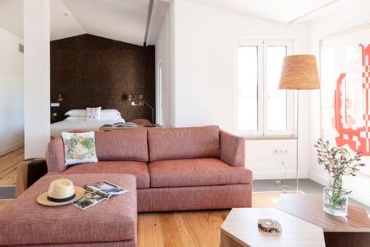 Une chambre d’hôtel moderne avec une grande fenêtre, un canapé rose, une table basse en bois et un lit doté d’une tête de lit sombre.
