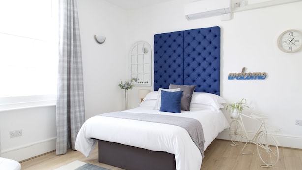 Un dormitorio moderno con un cabecero capitonado, cama con ropa de cama blanca, un banco gris, una mesita de noche blanca con una planta y una bicicleta.