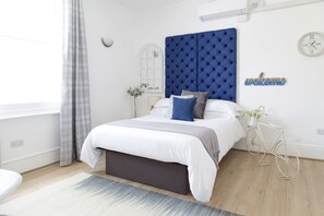 A modern bedroom with a tufted headboard, a bed with white bedding, a grey bench, a small white side table with a plant, and a bicycle.