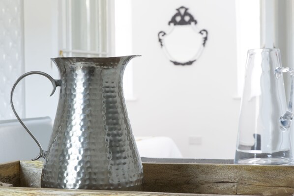 A metallic pitcher on a wooden tray with a glass pitcher in the background.