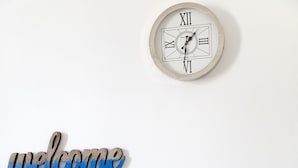 Pared blanca con un reloj, un letrero que dice 'welcome' y una bicicleta blanca con una cesta para plantas.