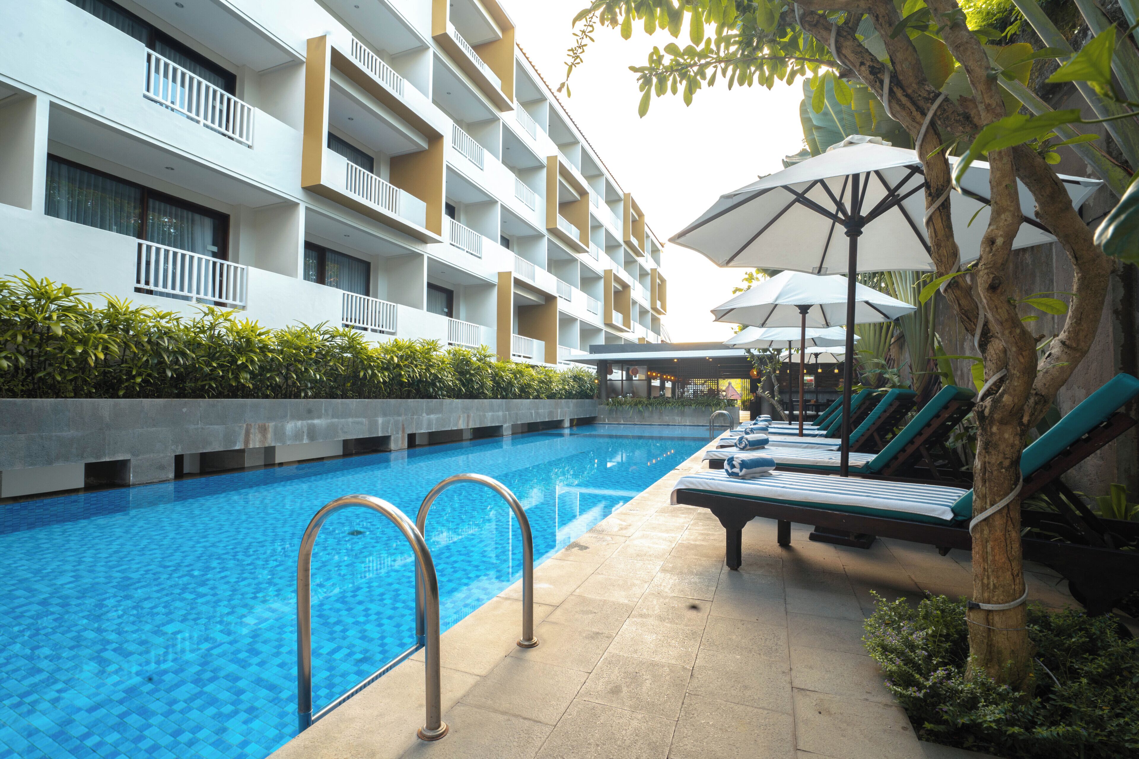Une piscine à l’eau d’un bleu cristallin, entourée de chaises longues et de parasols, et un immeuble à plusieurs étages doté de balcons.