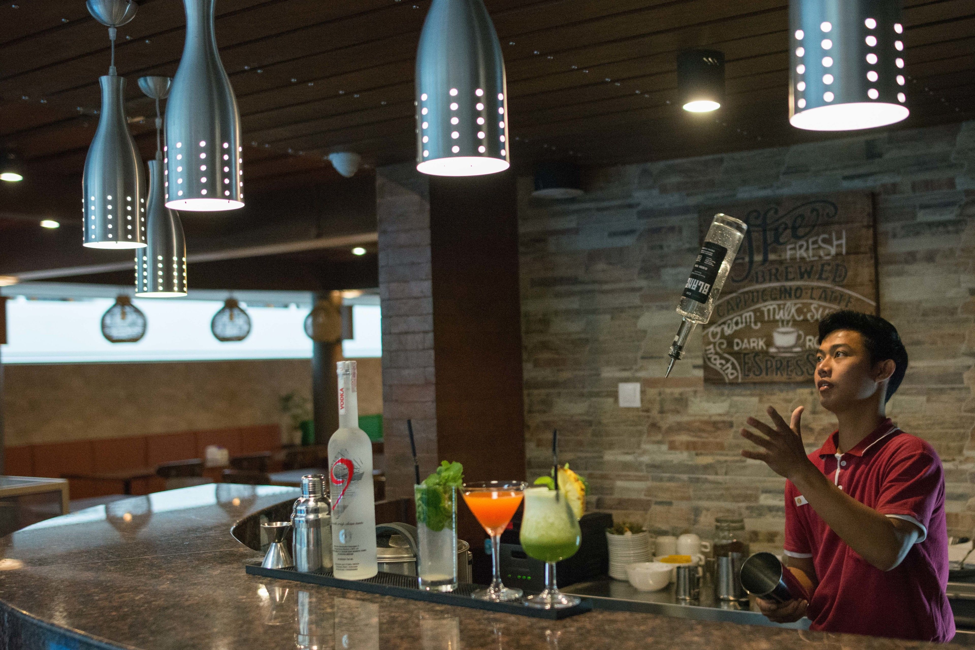 A bar area with hanging pendant lights, a counter with various drinks, and a wall with a coffee sign.