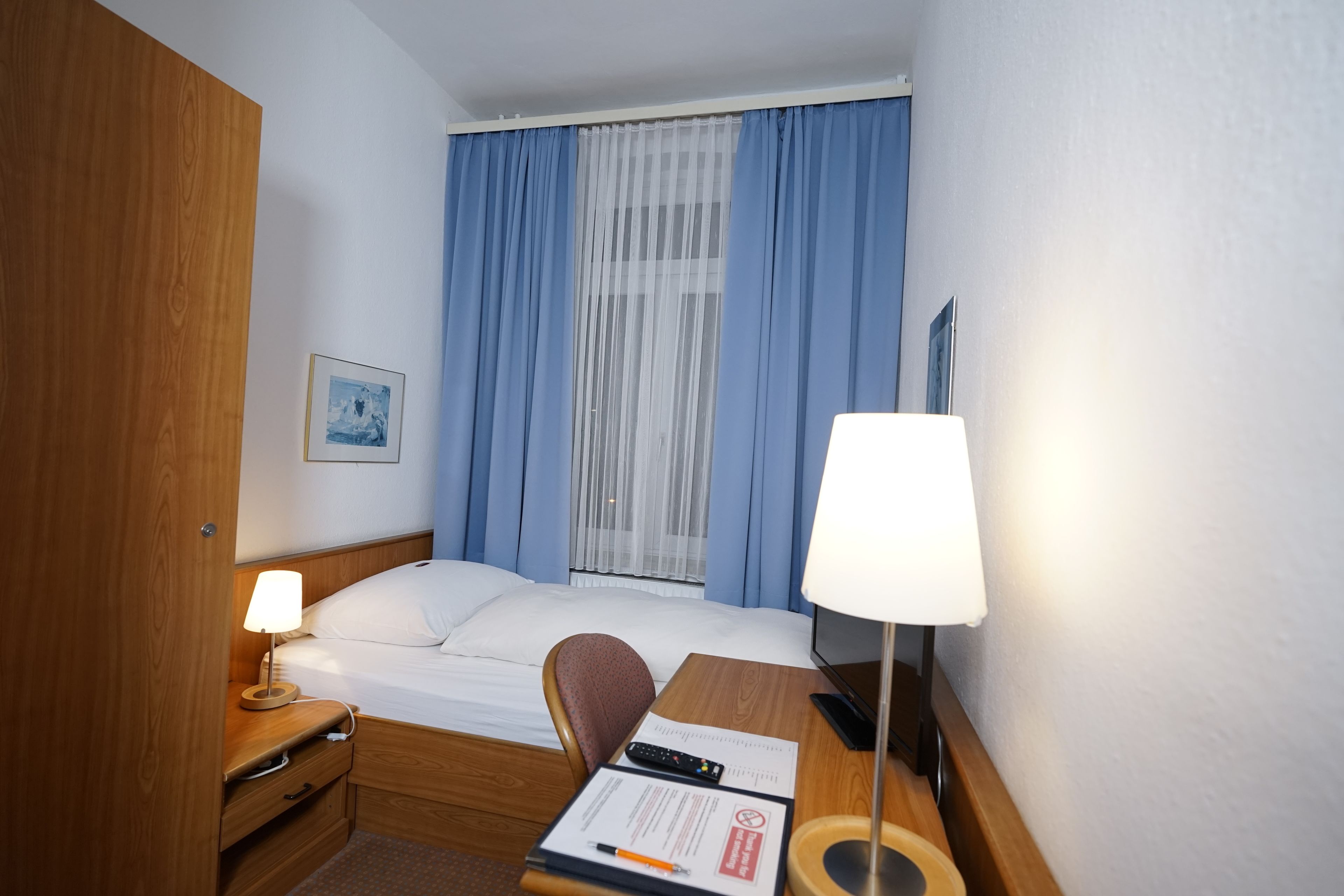 Habitación de hotel con cama, escritorio con lámpara, silla, televisor y ventana con cortinas.