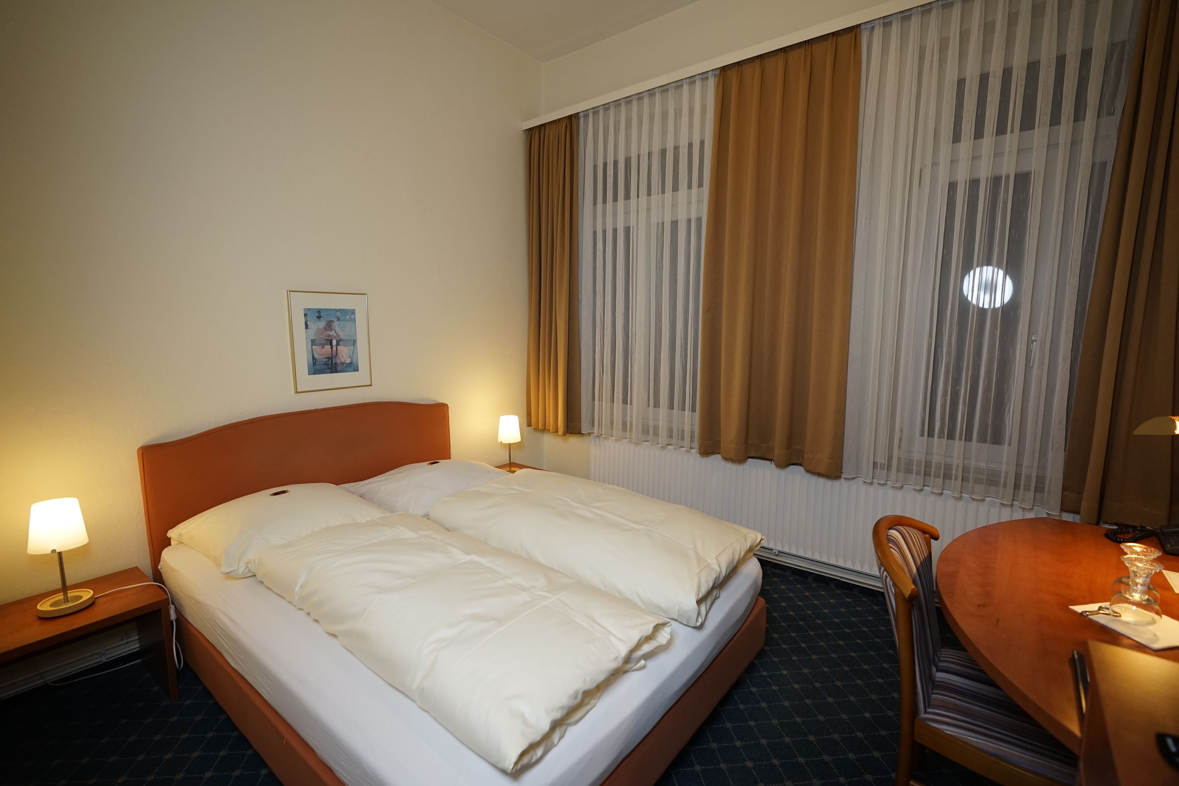 Habitación de hotel con cama, dos lámparas de noche, una silla de madera, una mesa de comedor y una ventana con cortinas.