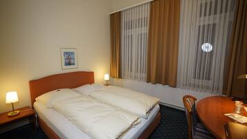 Habitación de hotel con cama, dos lámparas de noche, una silla de madera, una mesa de comedor y una ventana con cortinas.