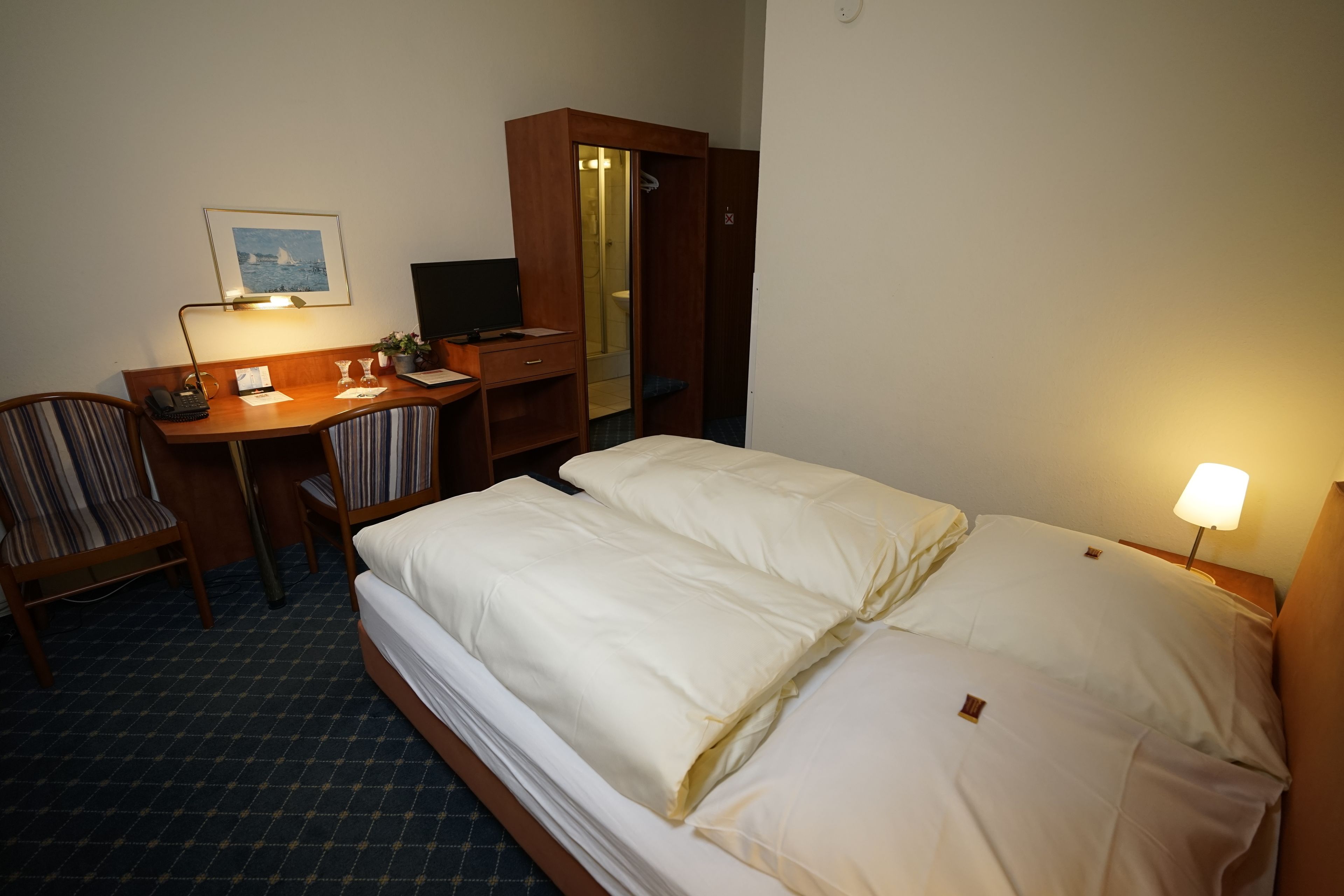 Una habitación de hotel con una cama, un escritorio, una silla, un armario y una lámpara de noche.
