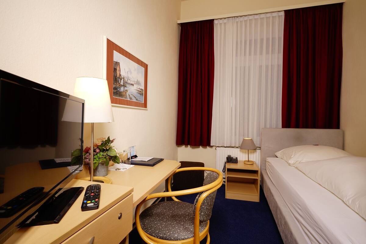 Une chambre d’hôtel avec un lit, un bureau avec un ordinateur, une chaise, une télévision et une fenêtre avec des rideaux.