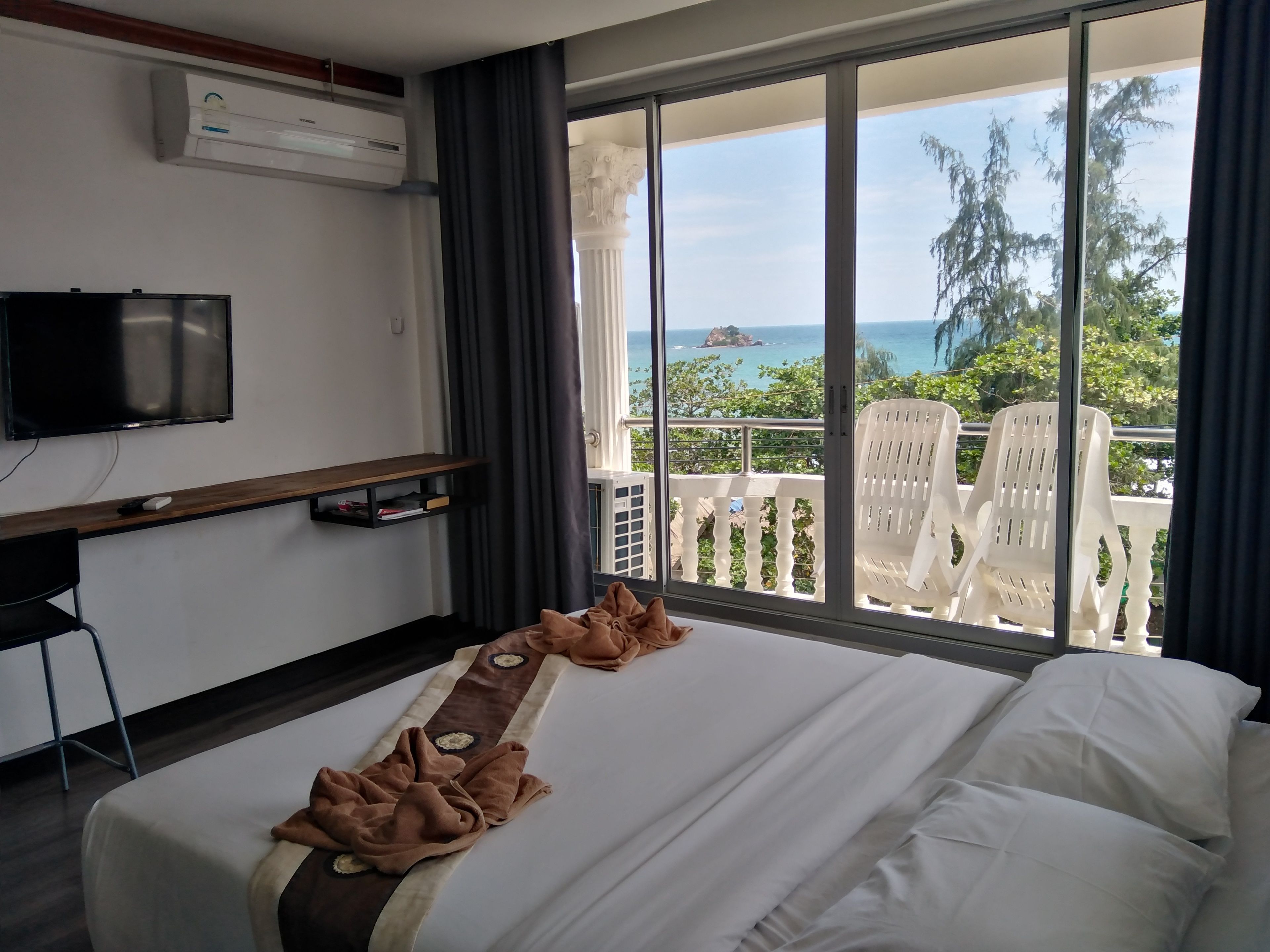 A hotel room with a bed, a TV, a desk with a computer, and a balcony with a view of the sea.