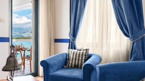 A blue sofa with a striped pillow, a wooden table with a bell, and a view of a balcony with a table and chairs. - Aparthotel Villa Lav (Tivat)