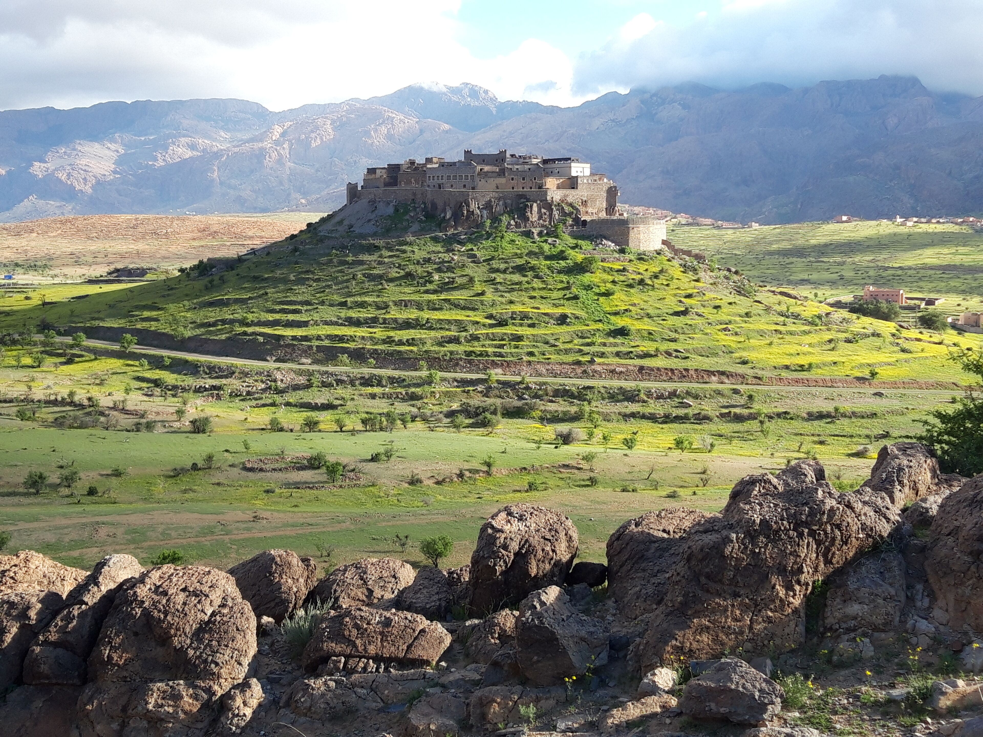 Une forteresse historique, perchée sur une colline verdoyante, avec des champs en terrasses et des montagnes en arrière-plan.
