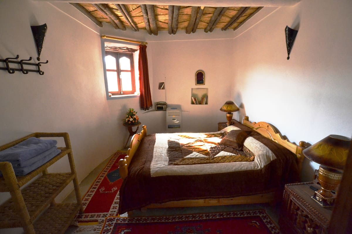 A bedroom with a wooden bed, a small table with a lamp, a window with curtains, a shelf with books, and a wall-mounted clock.