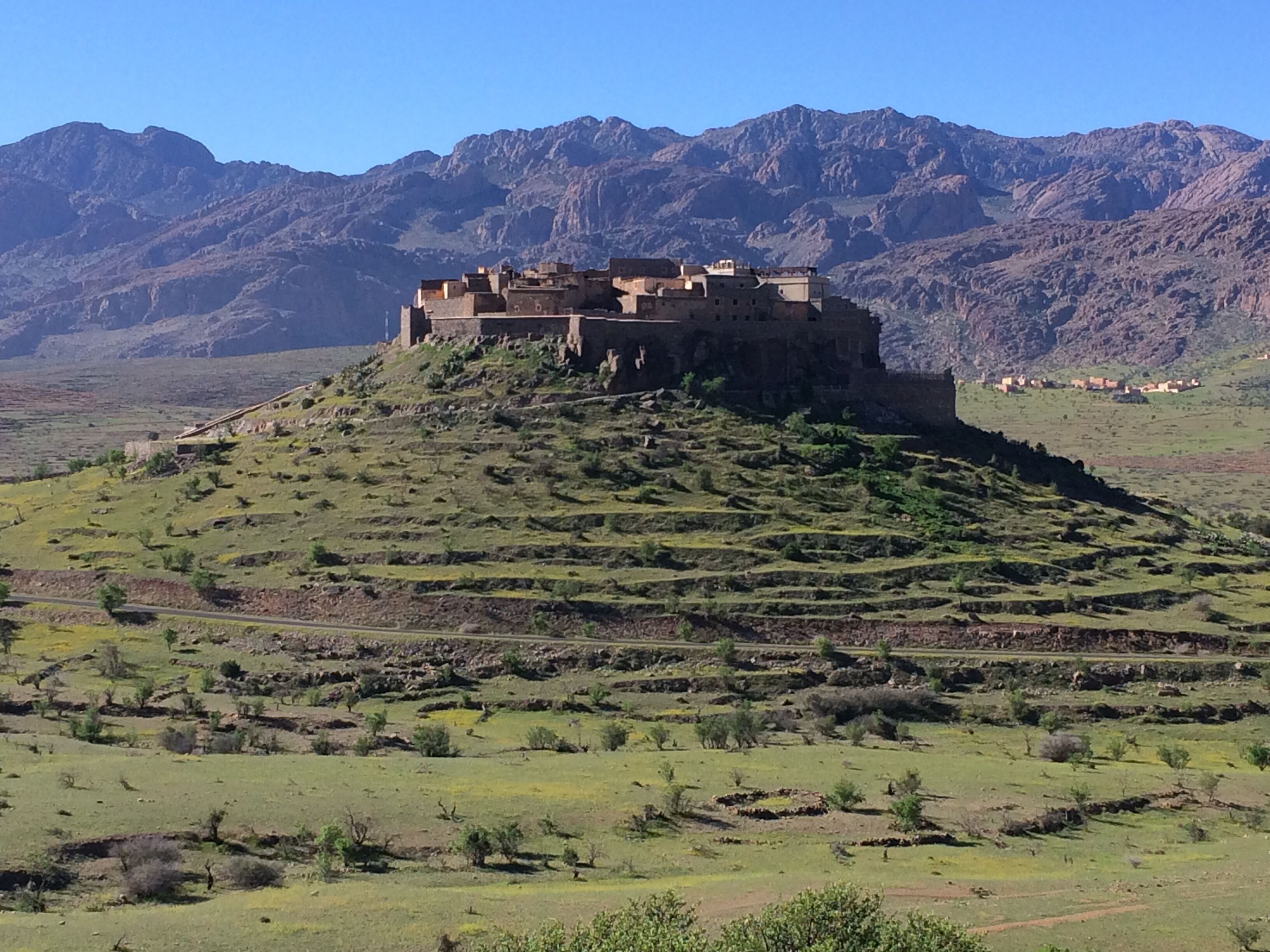 Un grand bâtiment fortifié, situé sur une colline, avec des champs en terrasses au premier plan et des montagnes en arrière-plan.