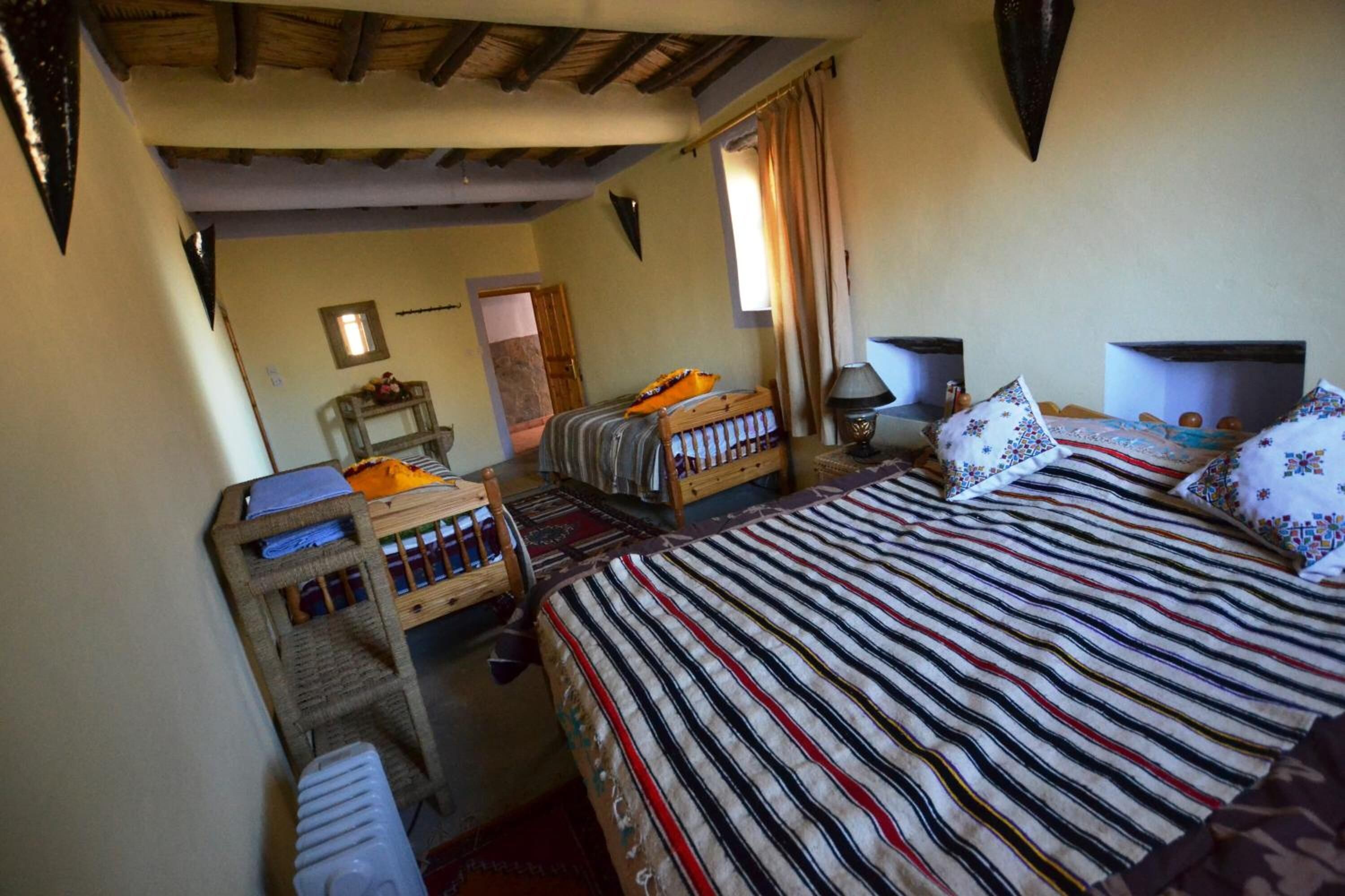 A cozy room with a bed, a small table, and a chair. There is a window with curtains, a radiator, and a wall-mounted shelf.
