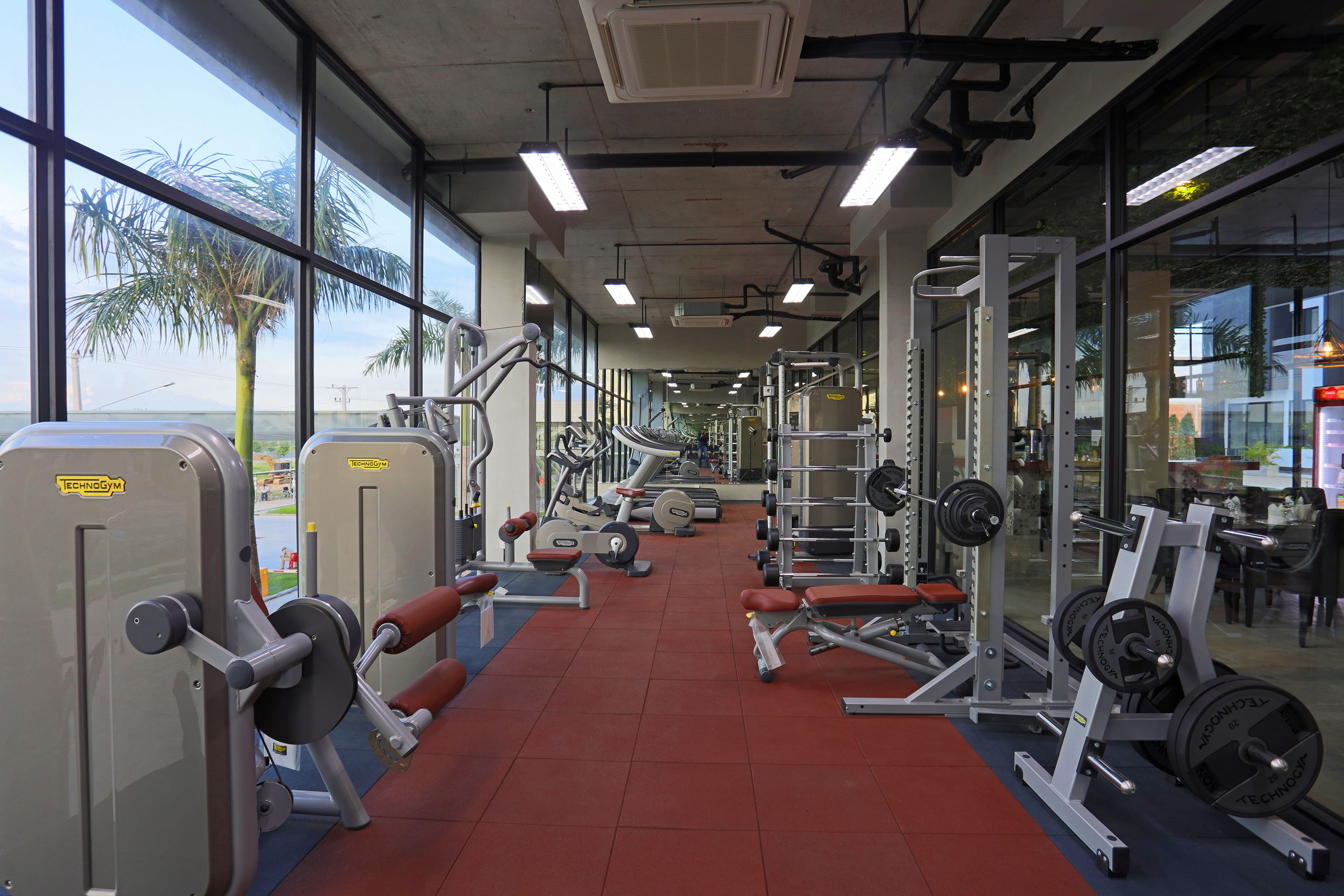Een sportschool met diverse fitnessapparaten, grote ramen en een rode vloer.