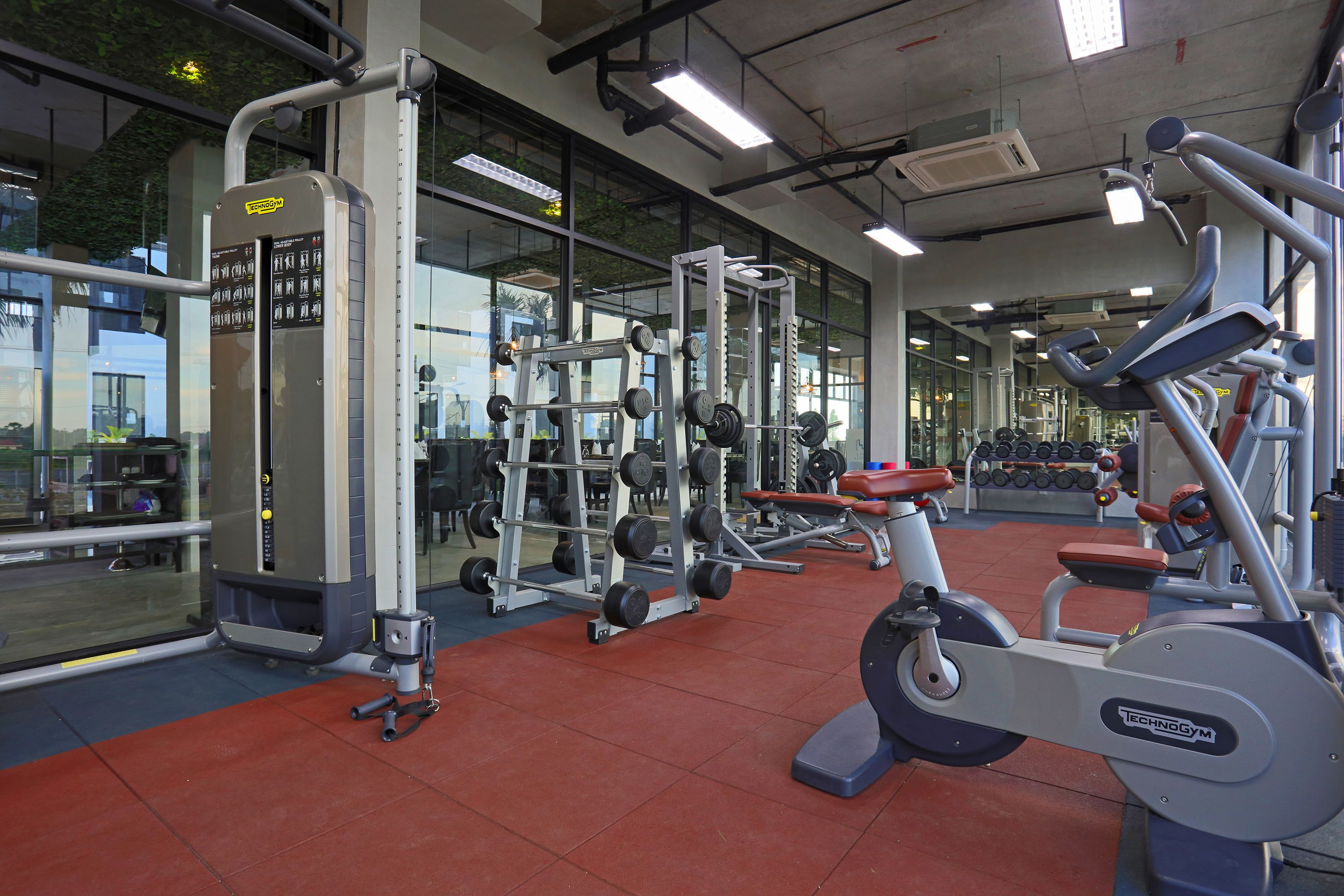 Un gimnasio con varios equipos de ejercicio, ventanas amplias y suelo rojo.