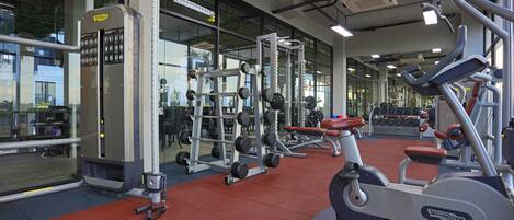 Un gimnasio con varios equipos de ejercicio, ventanas amplias y suelo rojo.