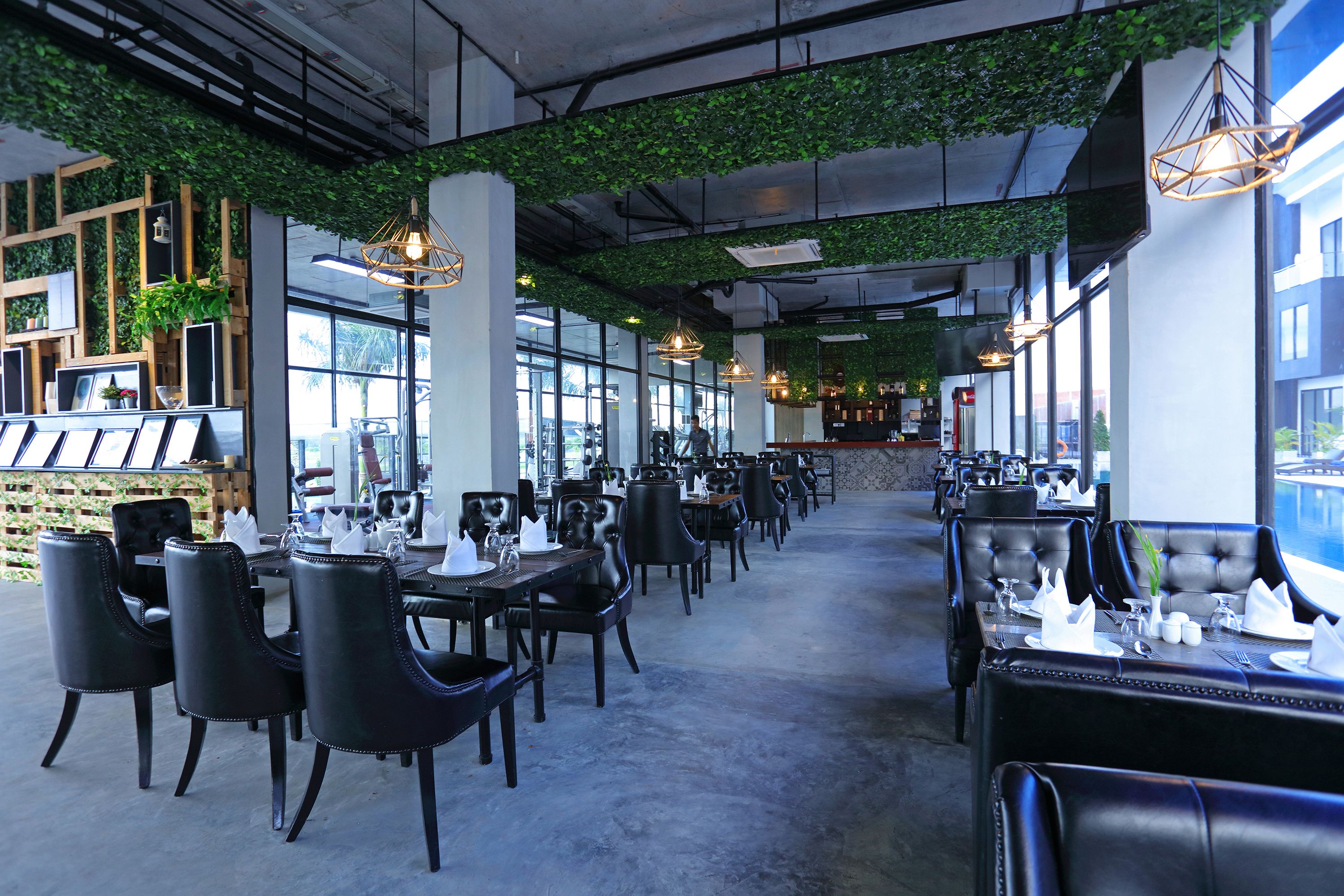 A spacious restaurant with high ceilings, green foliage, and modern pendant lighting. Tables are neatly set with white tablecloths and silverware.