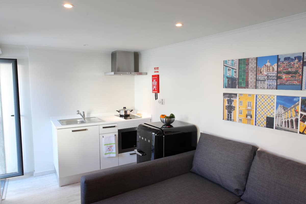 Cocina compacta con estufa moderna, un lavamanos y un refrigerador pequeño. En la zona de estar hay un sofá y una pared decorada con obras de arte coloridas.