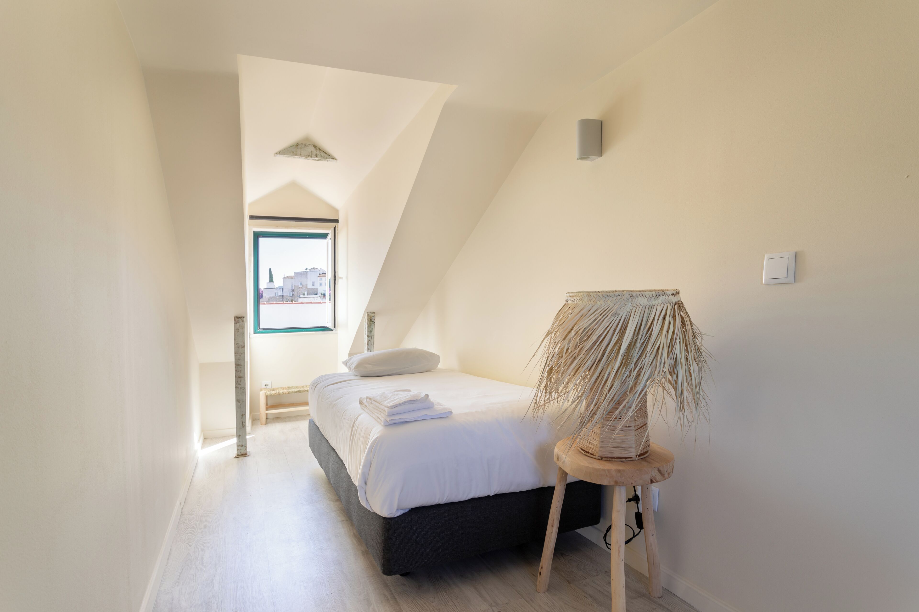 Ein Schlafzimmer mit Bett, Nachttisch, Lampe, Fenster und geneigter Decke.