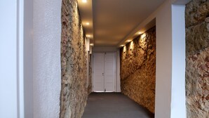 Um corredor estreito com paredes de pedra texturizadas e chão de madeira, que leva a uma porta branca no final.