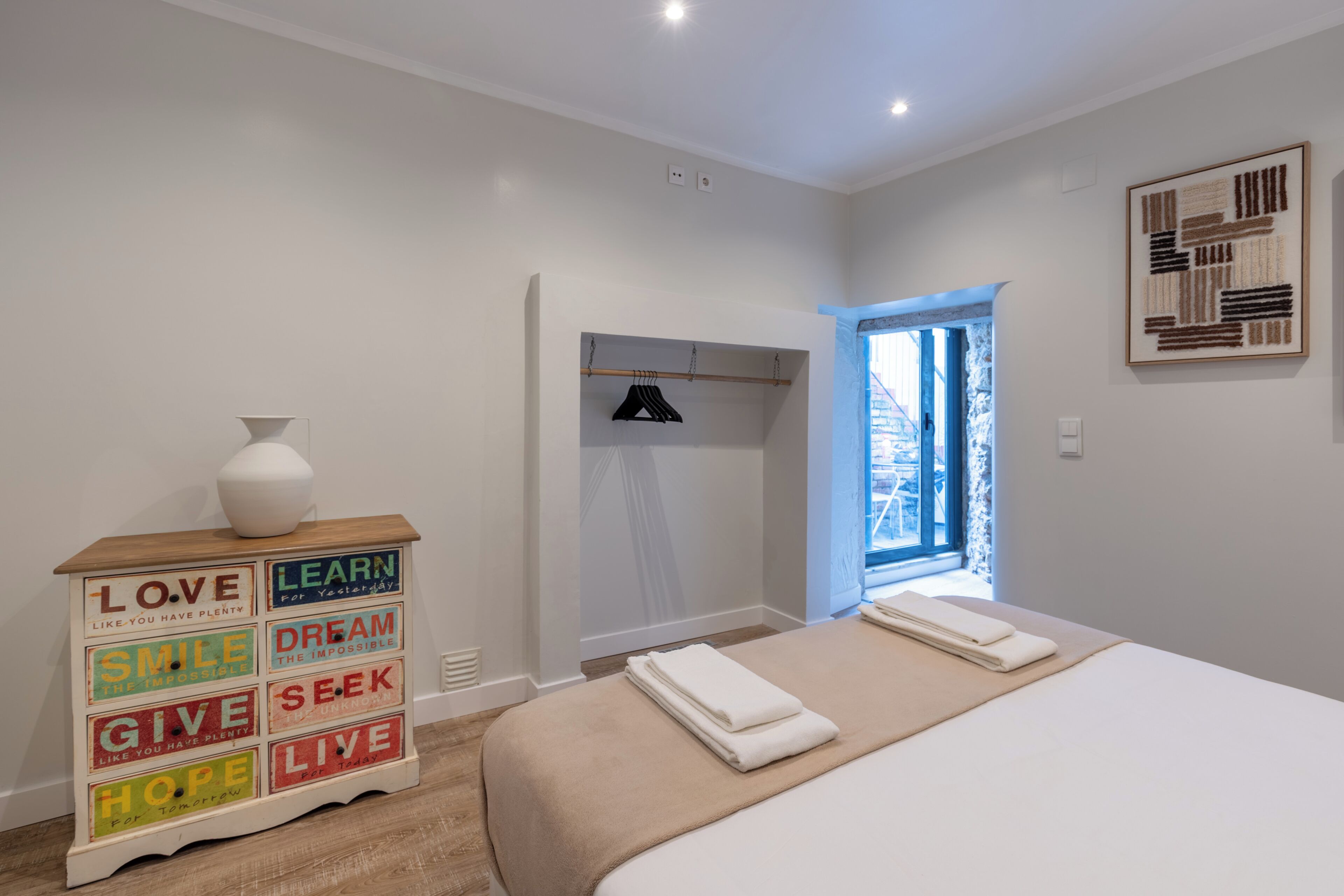 Un dormitorio con una cama, un aparador con palabras motivadoras, una ventana y una puerta.