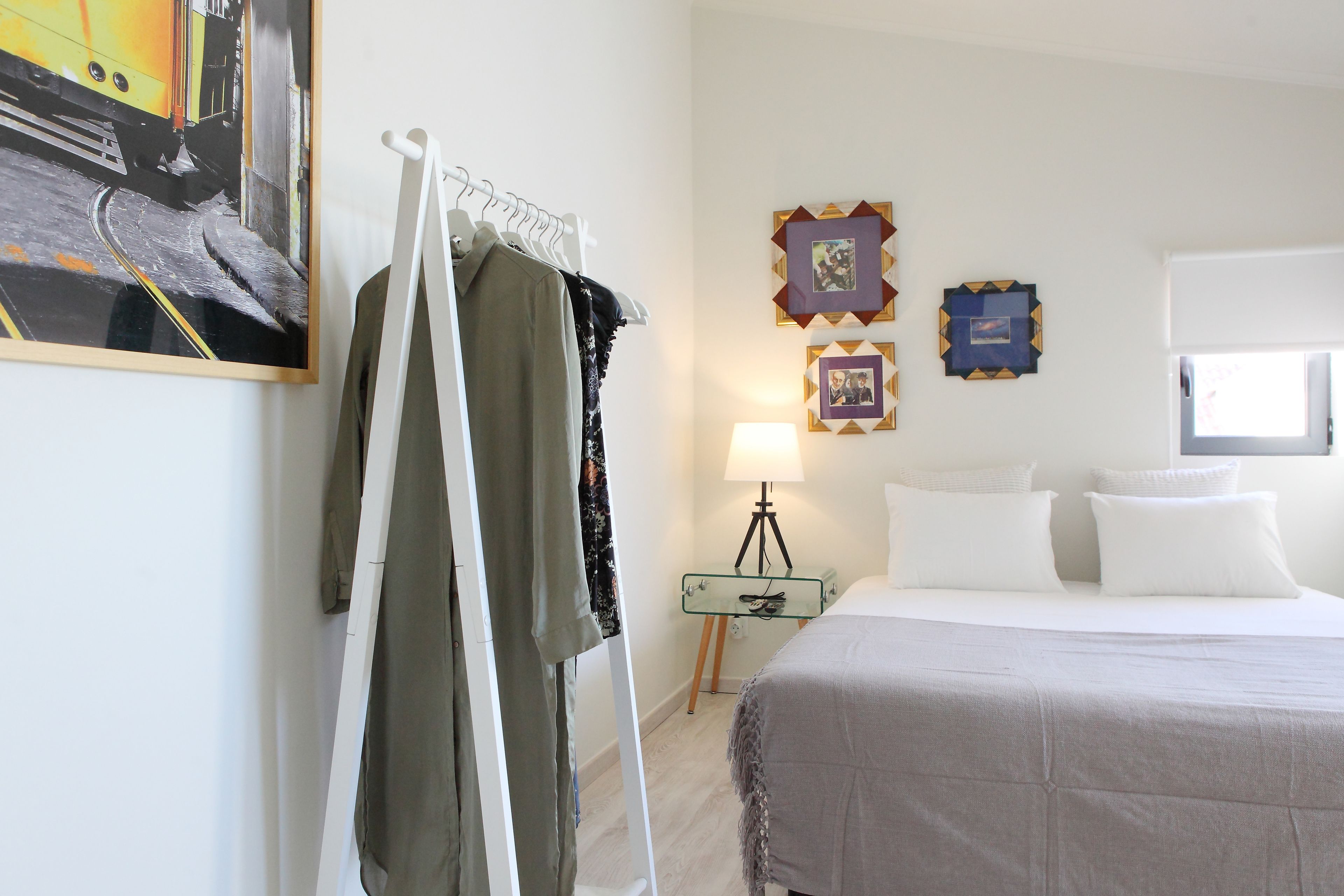 A hotel room with a bed, a clothing rack, a glass table with a lamp, and framed pictures on the wall.