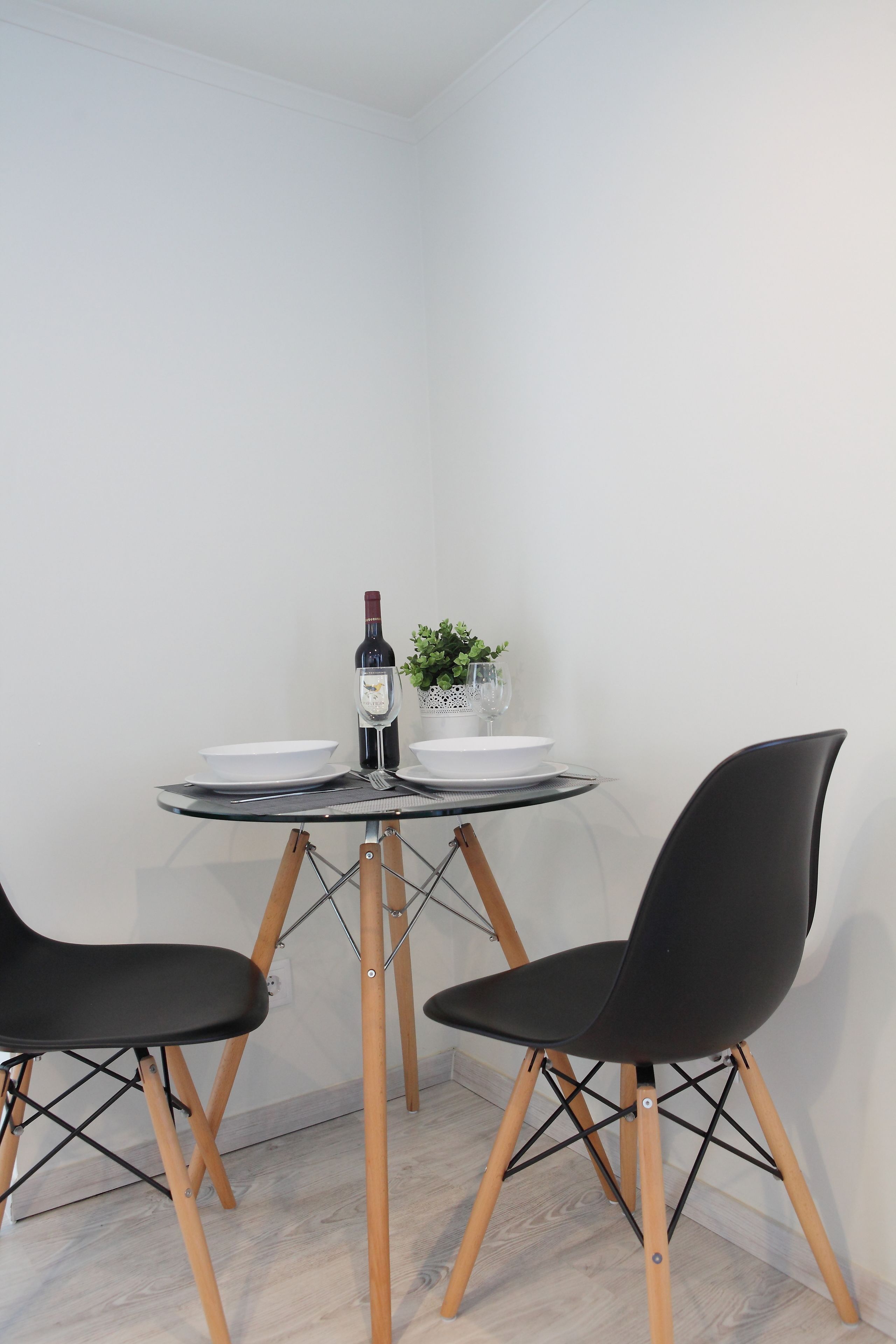 Een klein eetgedeelte met een glazen tafel, twee zwarte stoelen, een wijnfles en een kleine plant.