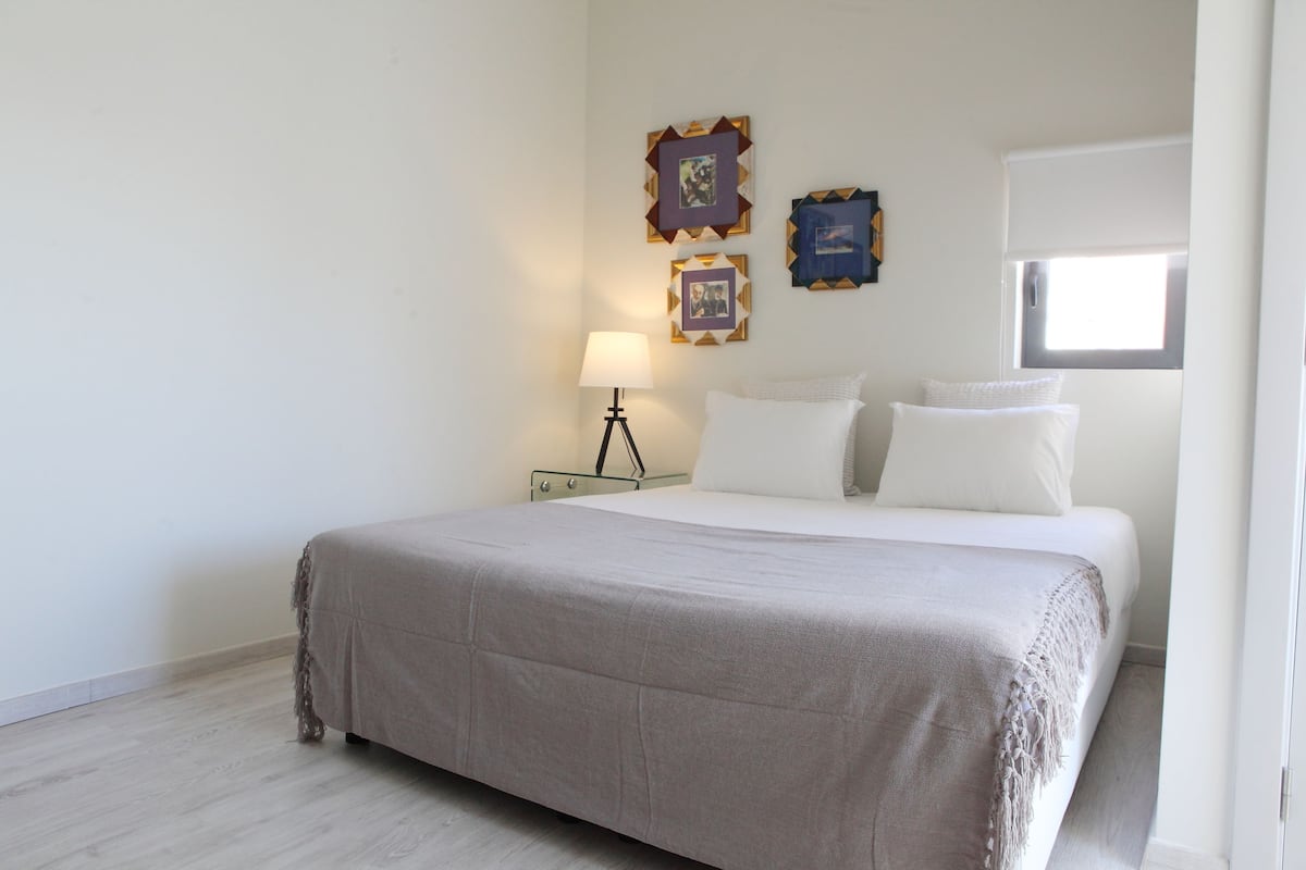 Una cama bien hecha con una colcha blanca y almohadas, una lámpara de noche y dos cuadros en la pared.