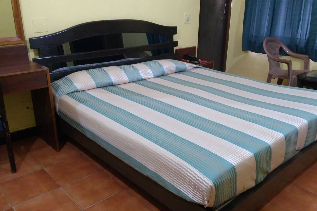 A single bed with a striped bedspread, a wooden headboard, a nightstand, a chair, and a small table with a lamp.
