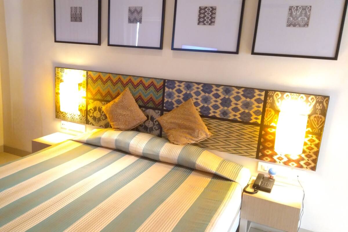 A hotel room with a bed, a chair, a bedside table with a phone, and framed artwork on the wall.