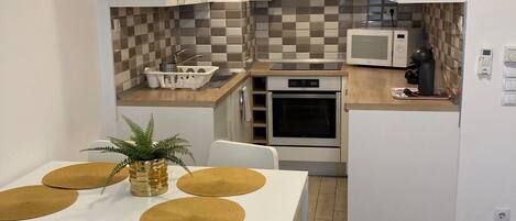 A compact kitchen with white cabinets, a microwave, an oven, and a sink. There is a dining table with white chairs and a plant on it.