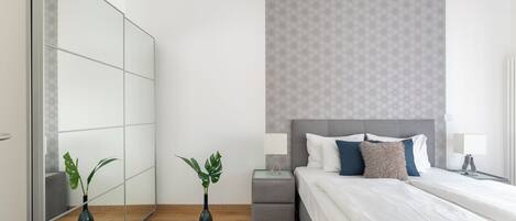 Une chambre moderne avec un grand miroir, une tête de lit grise, une table de chevet avec une lampe et un tapis moelleux.