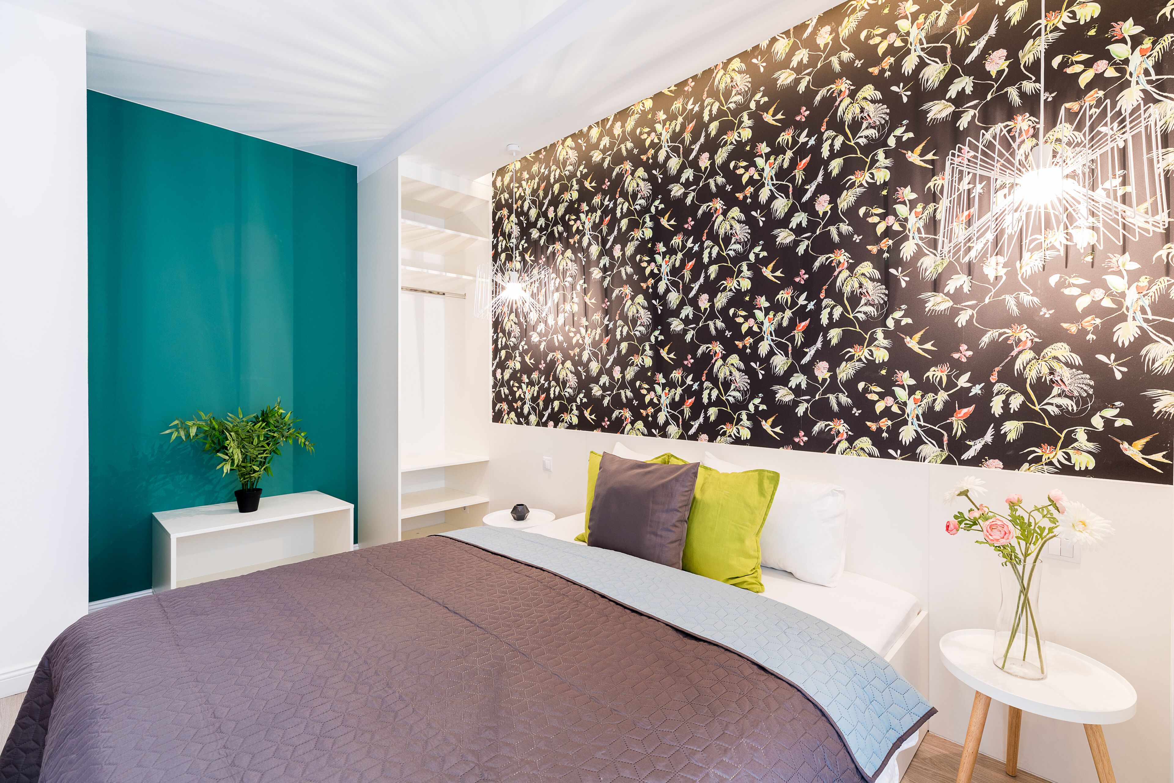 A modern bedroom with a large bed, a teal accent wall, a floral wallpaper, and a small table with a vase of flowers.