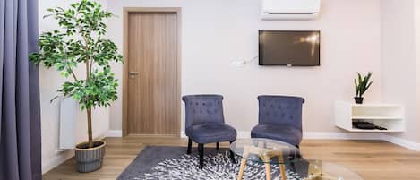 Una sala de estar moderna con una puerta de madera, dos sillones azules, una mesa de centro de vidrio, un tapete gris estampado, una planta en maceta y un televisor montado.